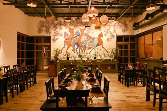 Interior of Maati Baani restaurant at Saj in the Forest with a vibrant Gond art mural, wicker pendant lights and wooden dining tables.