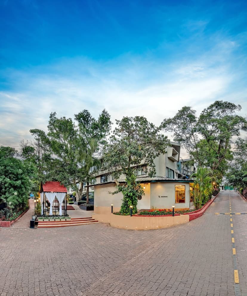 Wide-angle view of Saj On The Mountain resort facade with a paved driveway, garden gazebo and lush trees under a blue sky, Mahabaleshwar.