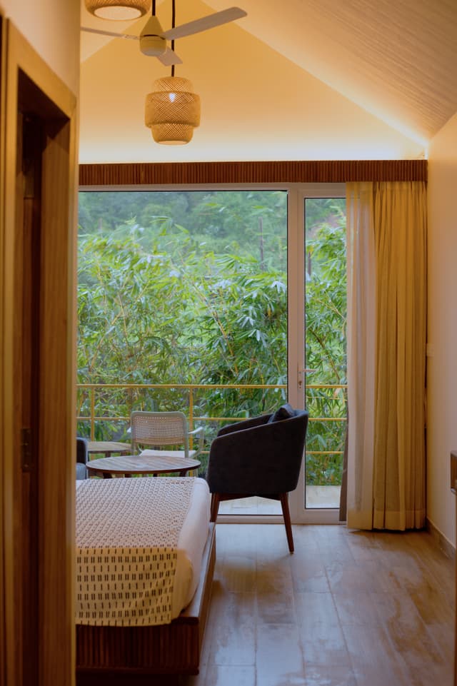 Cosy room interior at Saj By The Lake with a woven ceiling lamp and glass balcony doors opening to lush tree canopy views.