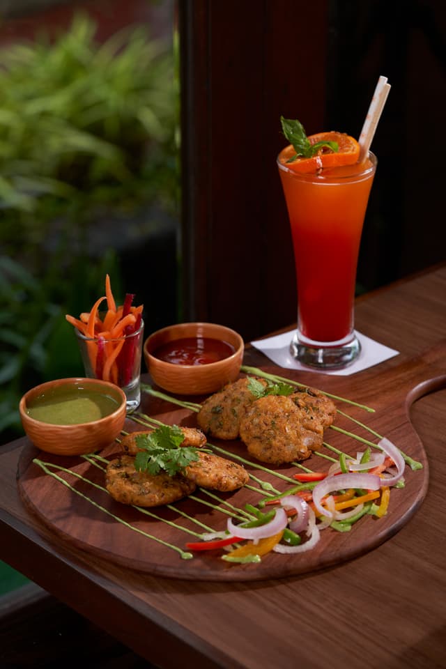 Crispy kebabs with chutneys and a fresh cocktail plated on a wooden serving board at Maati Baani restaurant, Saj By The Lake.